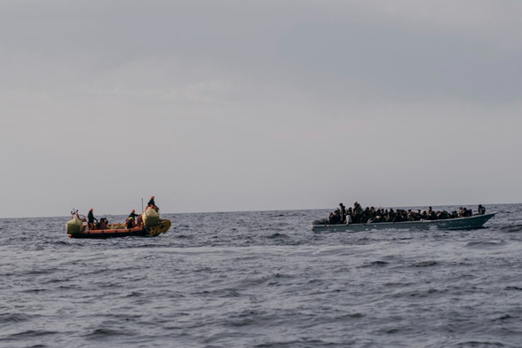 Rechts im Bild: Ein graublaues Holzboot treibt bei bedecktem Himmel auf dem Meer. Es ist voller Menschen. Niemand trägt eine Rettungsweste. Von links im Bild nähert sich sachte ein RHIB mit Seenotretter*innen. Sie haben mindestens zwei große Säcke mit Rettungswesten an Bord.