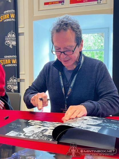 Une personne devant un livre avec des photos. La table est recouverte de rouge. Il fait face à une fenêtre. Il porte des lunettes ainsi qu'un pullover sombre. De son doigt, il pointe à une des photos du livre.
