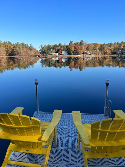Two Adirondack chairs at the end of a dock facing perfectly calm lake. The water reflects the blue sky