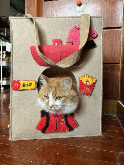 An orange and white cat looking out of a felt looking bag with McDonald’s patches on it