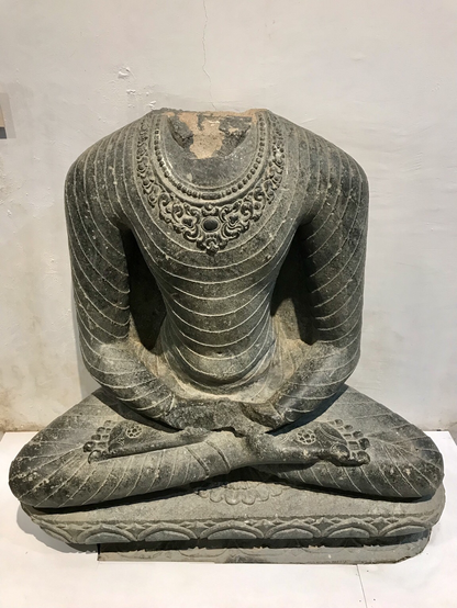 Buddha in Dhayan Mudra, 9. bis 10. Jahrhundert, Bodhgaya. Der kopflose Buddha hat die Beine über Kreuz mit den Fußsohlen nach oben zusammengeschlagen und seine Hände liegen vor dem Körper ineinander gelegt auf den Füßen. Sein Gewand scheint durchsichtig zu sein und hat eine verzierte Borte.
