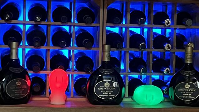 Ein Weinregal vor dem sich zwei LED-Leuchten in rot und grün befinden, die wie kleine Geister aussehen. Dazwischen drei Bocksbeutel-Flaschen.