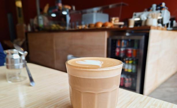 Picture of a glass with brown milky coffee in it, compact layer of foam (i.e. probably a Flat White) with heart latte art.The glass stands on a wooden surface, to the left a tea spoon leaning against a small metal and glass recepticle is visible as well. Blurred in tje backgroumd a counter with a beverage cooler is visible, with some confectionary or so, and a big coffee machine.
