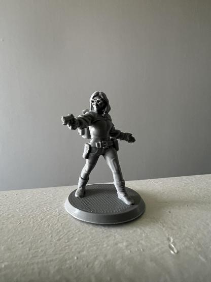 A gray miniature figure resembling a character in a tactical outfit, standing on a circular base with one arm extended as if holding a weapon. The background is plain and neutral-colored.