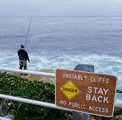 Fisherman on a cliff on a cloudy day, behind a fence with a sign saying Unstable Cliffs, Danger, Stay Back, No Public Access. It's a sheer cliff about 30 feet high in La Jolla, San Diego.
