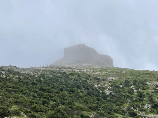 hill with green shrubs in the foreground, castle shrouded in clouds in the background