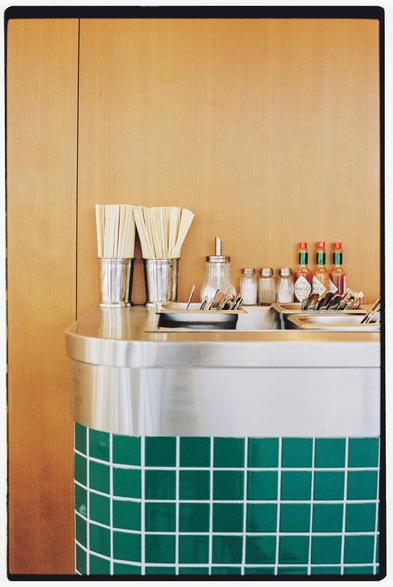 Photograph of the corner of a counter in a restaurant. Chopsticks, a sugar bowl, several salt shakers, and bottles of hot sauce are visible. The wall is wooden, the counter is metal, and the front of the counter is green and white tile.