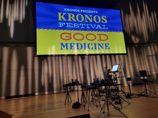 Photo of the stage and large  screen above it. The stage is empty of people but all the equipment is ready to go for the performers: portable music stands, tables, chairs, and tablet computers for the musical score. The screen shows what looks like the yellow and blue striped Ukrainian flag as its background. All text is block letters. At top is text Kronos Presents (white) Kronos (yellow) Festival (white). At bottom is text Good Medicine (good is red, medicine is blue). The fonts for each word vary a bit (e.g., serif vs. sans serif).

The stage is a light brown wood. The wall behind the screen is light brown vertical slats. It is illuminated with vertical white diffuse lights, so it looks almost like the pipes of an organ.
