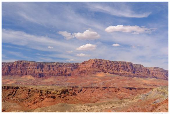 Panoramic view of the Vermillion Cliffs near Page, Arizona, showing layered red and orange rock formations rising above a dry, sparsely vegetated desert landscape under a blue sky with a few white clouds.
