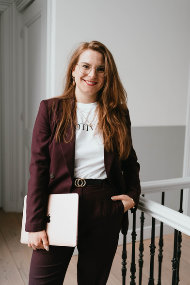 Woman in aubergine coloured suit leaning agains banister holding a laptop