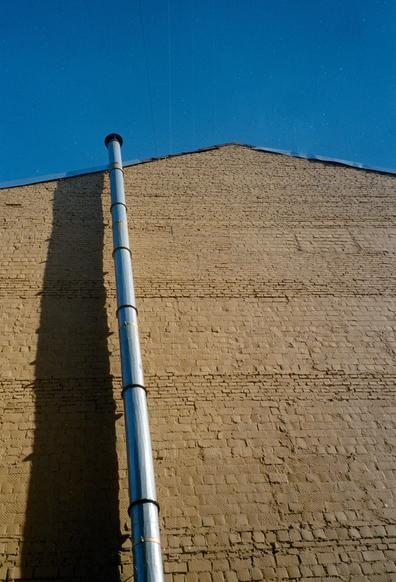 View from below of a brick, brown building with a triangular roof. The sky above the building is blue. Along the building, at the edge of the building is a metal pipe.