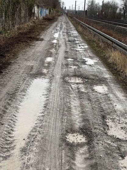 Ein schlammiger, unebener Feldweg erstreckt sich vor Ihnen, gesäumt von überwuchertem Gras und einer mit Graffiti bedeckten Wand auf einer Seite. Pfützen und Reifenspuren sind auf der Oberfläche sichtbar, mit Bahngleisen im Hintergrund.