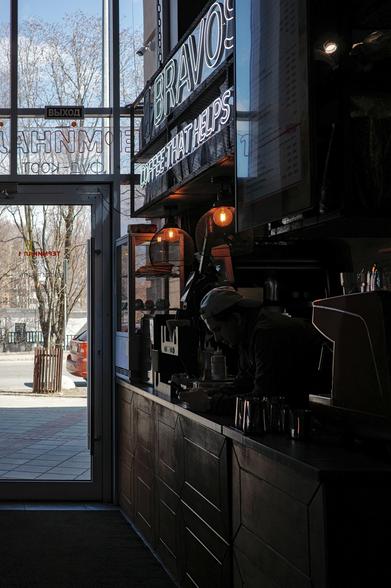 The barista is looking at his phone, leaning on the bar counter. It is dark in the cafe, light only from the street through the door and windows and from two lamps. 