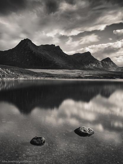 A photograph of Ben Loyal mountain reflected in the calm waters of Lochan Hakel in Sutherland, in the Scottish Highlands. The still surface of the loch creates a near-perfect mirror image of the mountain’s rugged peaks. Above, clouds drift gently across the sky, adding a soft, delicate layer to the monochrome scene and enhancing the sense of stillness.