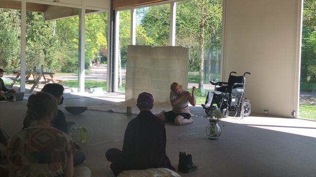 In this picture artist Toni Kritzer is sat on the floor in front of their wheelchair and a large white curtain that is hanging and partially draped on the floor, as part of the performance THE SICK GARDEN.