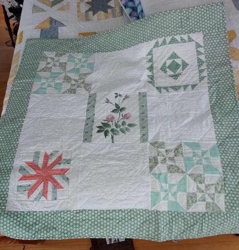 a quilt, it has a white and light green checkered thick border, inside is a white plane with 5 boxes, 4 in the corners and one in the center, each with a different geometric or floral design 