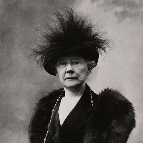 a photo of the artist, she is wearing furs and a large necklace, seen from her chest up, she is wearing a black hat with dark feathers sticking up out of it, she is looking at the viewer