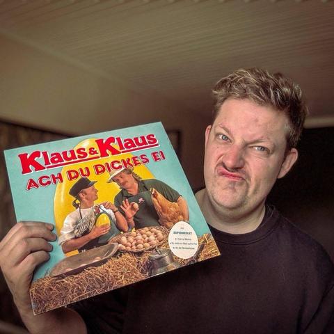 Eine Person hält eine bunte Vinylplatte mit dem Titel "Klaus & Klaus - Ach Du Dickes Ei" in der Hand. Auf dem Cover sind zwei Männer zu sehen, die in einer Bauernhofumgebung mit Hühnern und Eiern interagieren. Die Person hat einen verspielten, übertriebenen Gesichtsausdruck, der Humor oder Überraschung ausdrückt.