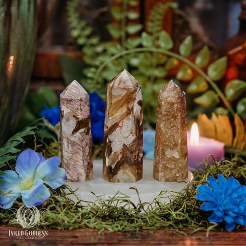 Three specimens of the "Golden Mica Tower" product are arranged on a selenite charging plate surrounded by moss and faux flowers. They are made of random patterns of white and brown with shimmering golden streaks. Presented by Inked Goddess Creations.