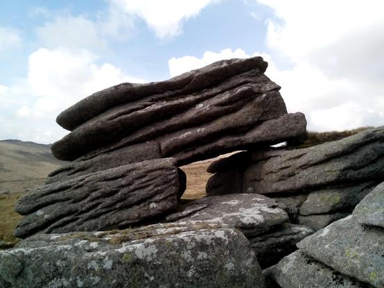 A close-up of a stack of granite that appears to be in layers tipping down to the left. In the centre is a gap through which the hillside beyond is visible.