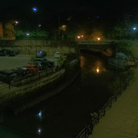 Photograph of the tidal stretch of Ouseburn River at south side of Ouseburn Bridge in Newcastle upon Tyne. To the left (south-west) of the bridge is Cut Bank, and to the right (north-east) is Byker Bank. In the foreground is the pedestrian Riverside Walk and the photo also spans some surrounding built-up areas and trees, with sky in the background. Still image captured from webcam at 03:12 hrs on Tuesday 29 April 2025.