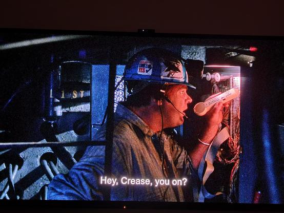 Poorly lit nighttime photo of a tv screen showing a scene from the movie Sneakers. Dan Akroyd as Mother with hard hat and headset microphone, is in a cable vault testing phone lines with a handheld device. Subtitle reads "Hey, Crease, you on?"