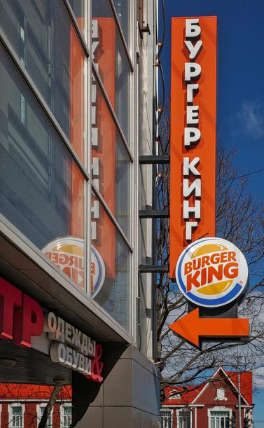 A large street-side, red-orange Burger King sign is reflected in the windows of a neighboring building. Behind the sign you can see a red house and blue skies.