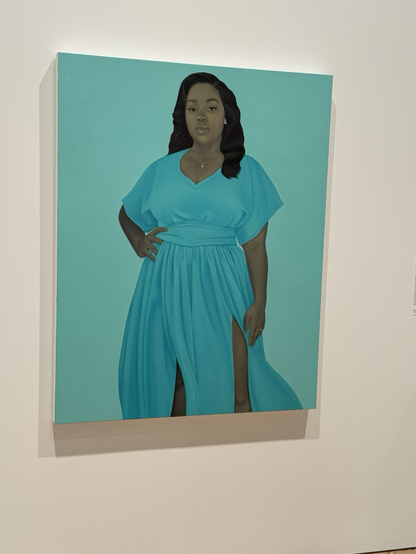 A close-up of Amy Sherrald’s portrait of Breonna Taylor—she gazes at the viewer, with one hand at her waist. She has shoulder-length black hair, a flowing turquoise dress. The background is lighter turquoise blue