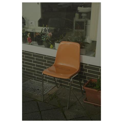 Ein alter orange farbener Platikschslen-Stuhl steht auf der Straße vor einem Schaufenster eines Friseursalons. Ein Pfütze mit Regenwasser hat sich auf seiner Sitzfläche gebildet. 