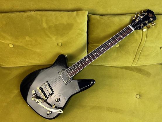 Eine schwarze Retro-E-Gitarre auf einem grün/goldenen Samtsofa. 