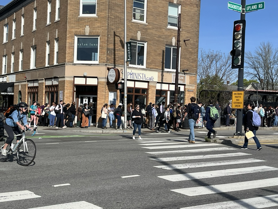 A long line of people waiting outside a three-story brick building on a corner near crosswalk in Cambridge Massachusetts on mass Avenue/Porter Square