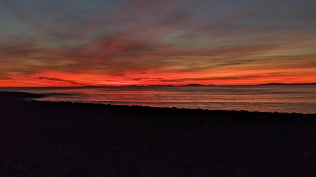 Historical Documentation in the form of a photograph of a rising sun over the ocean bay at Yirrell Beach in Winthrop, MA. The sky is filled with vibrant orange and red hues, with the sun rising in the distant horizon while casting a warm glow over the water. The water is calm and reflects the colors of the sky. In the foreground, there is a rocky shoreline with small waves crashing onto the shore. The horizon line is visible in the distance, with a few small islands scattered across it. The overall mood of the image is peaceful and serene.