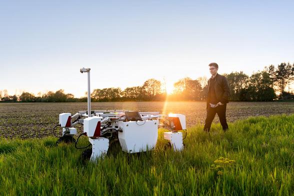 On voit un paysage au couché du soleil. Un homme marche derrière un robot agricole qui désherbe un champ.