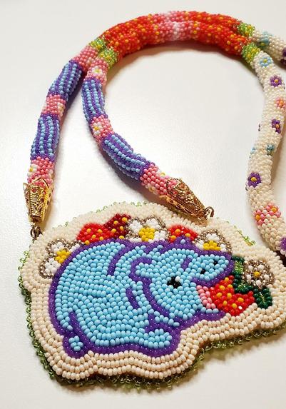 a beaded medallion made by kiamichi of a baby hippo in blue eating a strawberry in a field of strawberries and their flowers. the necklace that holds the medallion is multiple colors with floral accents. 