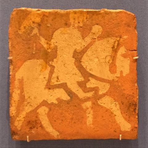 An orangey coloured square tile with the design of a falconer picked out in a paler shade.
