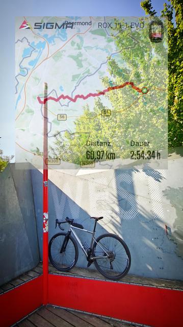 Ein Gravelbike steht auf einem roten Podest an eine graue Wand gelehnt. Auf der Wand befindet sich ein großer weißer Schriftzug WEST. Links ein roter Mast. Oben im Bild ist eine Landkarte mit dem Streckenverlauf eingeblendet. Ausserdem die Distanz 60,97km und die dafür benötigte Zeit 2:54:34h Und der Himmel ist blau.