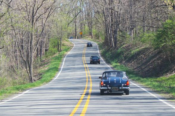 Four dark-colored MGBs driving uphill on a winding country road through the woods. The grasses and shrubs on the side are green while the trees are just starting to bud.