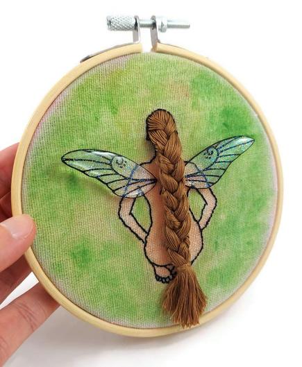 Broderie d'une fée nue assise de dos, avec une longue tresse et des ailes en relief.
Elle est réalisée sur une toile en lin peinte en vert, et fixée à un cercle à broder rond en bois clair.
Les ailes de la fée sont réalisées en CD recyclé, découpées et peintes à la main, puis recouvertes de paillettes et de résine époxy.