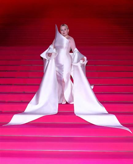 Nicola Coughlan on the pink carpet on stairs in her beautiful white outfit. Formfitting satin white dress with the right shoulder sculpted upwards into a tip and a white cape/shawl so that she has bare shoulders but sleeves from the ellbow down. The sleeves fall down and become a train right an left in front of her. She wears white pumps and her blond hair is slicked back into a chignon.
