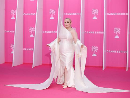 Nicola Coughlan on the pink carpet in her beautiful white outfit. Formfitting satin white dress with the right shoulder sculpted upwards into a tip and a white cape/shawl so that she has bare shoulders but sleeves from the ellbow down. The sleeves fall down and become a train right an left in front of her. She wears white pumps and her blond hair is slicked back into a chignon.