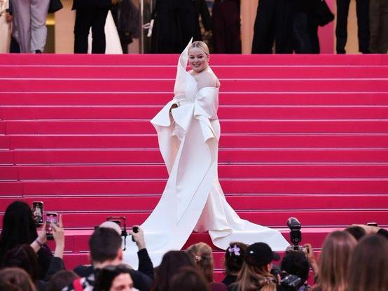 Nicola Coughlan on the pink carpet on stairs in her beautiful white outfit, photographed from the side mid turn. Formfitting satin white dress with the right shoulder sculpted upwards into a tip and a white cape/shawl so that she has bare shoulders but sleeves from the ellbow down. The sleeves fall down and become a train right an left in front of her. She wears white pumps and her blond hair is slicked back into a chignon.