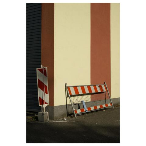 Vor einer beige-rot gestreiften Wand lehnt ein rot gestreiftes Baustellen Absperrgitter. 