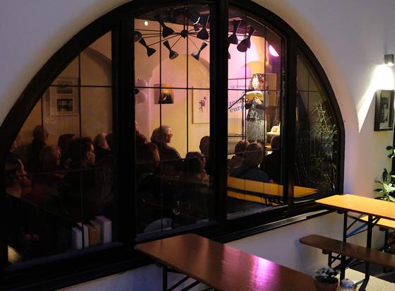 Eine Außenimpression der Lesung, durchs Fenster im Innenhof des Aegis Cafés fotografiert. Fee liest stehend am Mikrofon, gut sichtbar auch das gebannte Publikum.