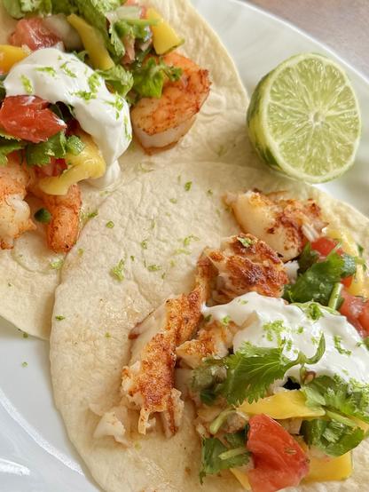 A closeup of two tacos overlapping, one with spiced shrimp, one with spiced fish, both with mango salsa, and a dollop of lime crema. Half of a lime sits between.  On a  round, white plate.