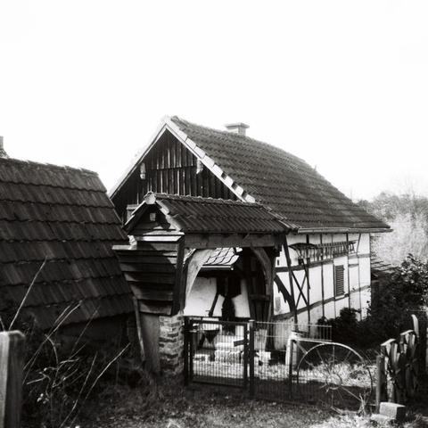Black and white photo of a small half-timbered farmhouse with a gate heading into an inner courtyard.