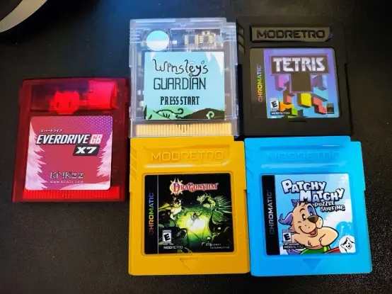 An image of five Game Boy cartridges.

A transparent red Christmas edition of the Everdrive GB x7, which has white pine trees on it's label.
A transparent white cartridge for Winsley's Guardian.
A black cartridge for Tetris.
A gold (maybe spicy mustard) cartridge for Dragonyhm.
A bright blue cartridge for Patchy Matchy.