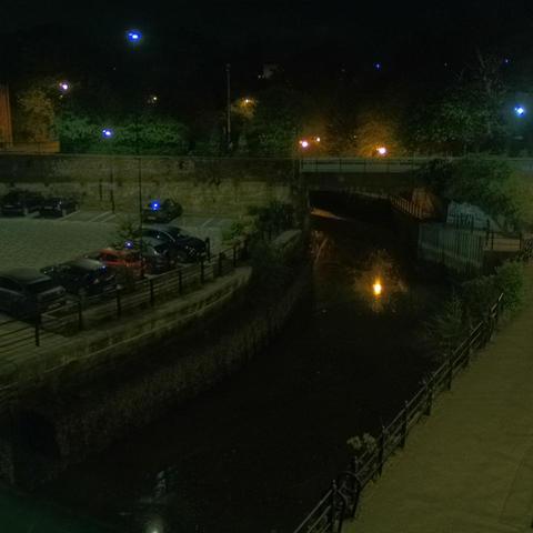 Photograph of the tidal stretch of Ouseburn River at south side of Ouseburn Bridge in Newcastle upon Tyne. To the left (south-west) of the bridge is Cut Bank, and to the right (north-east) is Byker Bank. In the foreground is the pedestrian Riverside Walk and the photo also spans some surrounding built-up areas and trees, with sky in the background. Still image captured from webcam at 03:12 hrs on Wednesday 30 April 2025.
