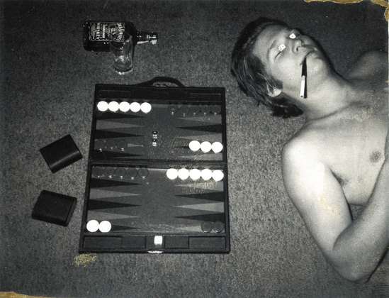 Black & white photograph from the 1970's of Jeff Bridges lying down shirtless with a cigarette with one of those black plastic mouthpieces at the end of it and a pair of dice laying over his closed eyes. To his left is a backgammon board with the pieces in play. In the background is a fifth of Jack Daniels laying on its side and a tall cocktail glass. 
