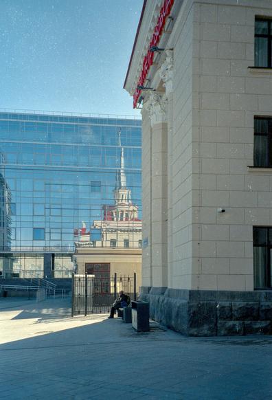 In the foreground, in the shadow, part of the building of the railway station. A man is sitting at the station. In the far background, sunlit, glazed wall of a modern building, which reflects the building of the station. 