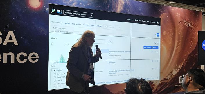 Man speaks in front of large screen on which search results from Science Explorer can be seen.  NASA Science logo is partially visible on the left.